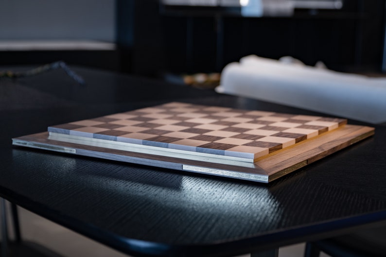 May include: A wooden chessboard with a light brown and dark brown chequered pattern. The board is closed and sitting on a black table.