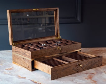 Könnte beinhalten: Eine Holzuhrbox mit Glasdeckel und einer herausziehbaren Schublade. Die Box hat eine dunkelbraune Oberfläche und ist aus Walnussholz gefertigt. Die Box hat mehrere Fächer zur Aufbewahrung von Uhren.