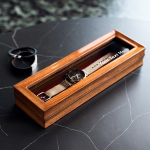 Puede incluir: Una caja de madera para relojes con tapa de cristal y un reloj plateado en su interior. La caja tiene el texto "Your Text Here" grabado en la parte superior.