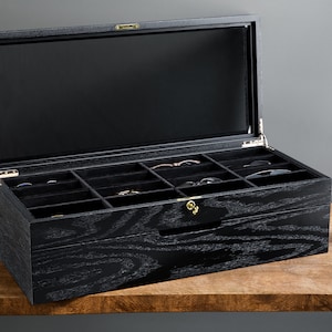 May include: Black wooden box with a glass lid and a drawer. The box has multiple compartments for storing sunglasses. The box is open and the compartments are empty.