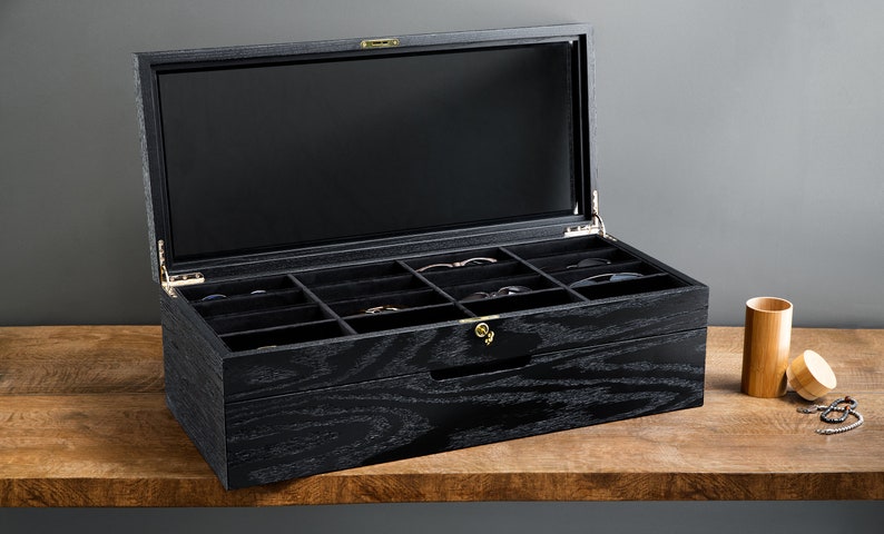 May include: Black wooden box with a glass lid and a drawer. The box has multiple compartments for storing sunglasses. The box is open and the compartments are empty.