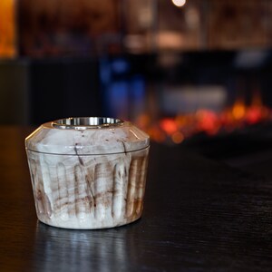 May include: A small, round, wooden container with a silver metal top. The container has a light brown and white marbled pattern. It is sitting on a dark wooden surface.