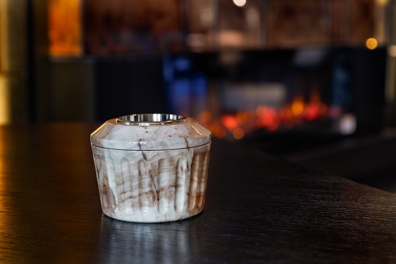 May include: A small, round, wooden container with a silver metal top. The container has a light brown and white marbled pattern. It is sitting on a dark wooden surface.