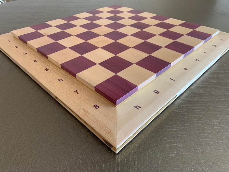 Puede incluir: Un tablero de ajedrez de madera con un marco marr&oacute;n claro y un patr&oacute;n de tablero de ajedrez marr&oacute;n claro y morado. El tablero est&aacute; etiquetado con los n&uacute;meros del 1 al 8 y las letras de la a a la h.