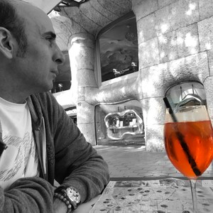 May include: A man sits at a table outside a building, looking off to the side. A glass of orange juice with a straw is on the table in front of him.