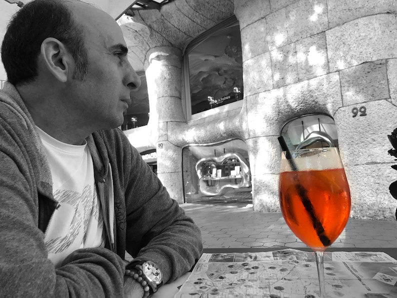 May include: A man sits at a table outside a building, looking off to the side. A glass of orange juice with a straw is on the table in front of him.