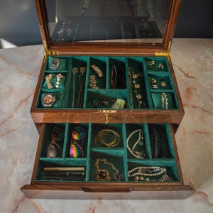 Może przedstawiać: Drewniane pudełko na biżuterię ze szklanym blatem i wieloma przegródkami wyłożonymi zielonym aksamitem. Pudełko jest wypełnione różnymi elementami biżuterii, w tym zegarkami, kolczykami, naszyjnikami, bransoletkami i okularami przeciwsłonecznymi. Pudełko jest otwarte, ukazując zawartość.