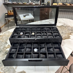 Może przedstawiać: Czarne drewniane pudełko na zegarki ze szklanym wierzchem i szufladą. Pudełko ma 36 przegródek do przechowywania zegarków. Szuflada ma 36 przegródek do przechowywania zegarków.
