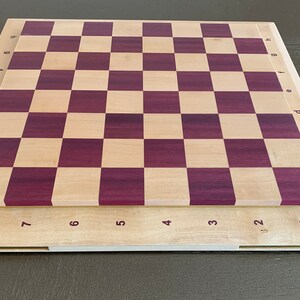 Puede incluir: Un tablero de ajedrez de madera con un acabado en madera natural y morado. El tablero tiene n&uacute;meros del 1 al 8 en los lados y letras de la a a la h en los extremos.