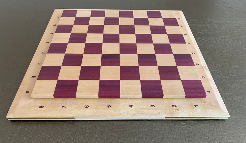 Puede incluir: Un tablero de ajedrez de madera con un acabado en madera natural y morado. El tablero tiene n&uacute;meros del 1 al 8 en los lados y letras de la a a la h en los extremos.