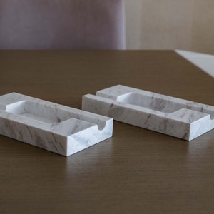Op de afbeelding: Twee witte marmeren telefoonstandaards met een rechthoekige vorm en een groef om een telefoon vast te houden. De standaards staan op een houten oppervlak.