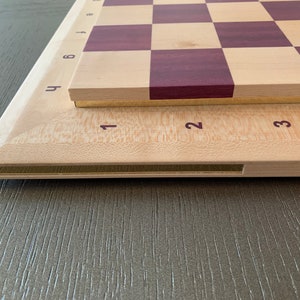 Puede incluir: Un tablero de ajedrez de madera con una superficie de madera clara y cuadrados de color morado oscuro. El tablero tiene un borde de lat&oacute;n y est&aacute; numerado del 1 al 8 en el lateral. El tablero est&aacute; sobre una superficie de madera.