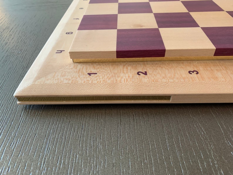 Puede incluir: Un tablero de ajedrez de madera con una superficie de madera clara y cuadrados de color morado oscuro. El tablero tiene un borde de lat&oacute;n y est&aacute; numerado del 1 al 8 en el lateral. El tablero est&aacute; sobre una superficie de madera.