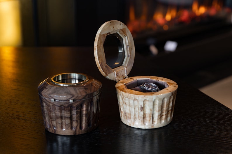 May include: Two wooden watch boxes, one open with a watch inside and the other closed. The boxes are made of light and dark wood with a decorative pattern.