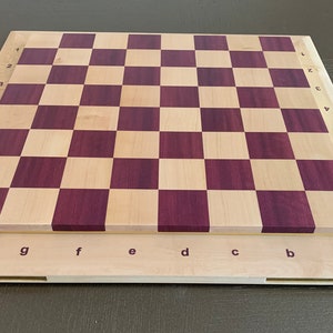 Puede incluir: Un tablero de ajedrez de madera con un patr&oacute;n de tablero de ajedrez de madera clara y madera p&uacute;rpura. El tablero tiene n&uacute;meros del 1 al 8 en el lateral y letras de la a a la h en la parte inferior.