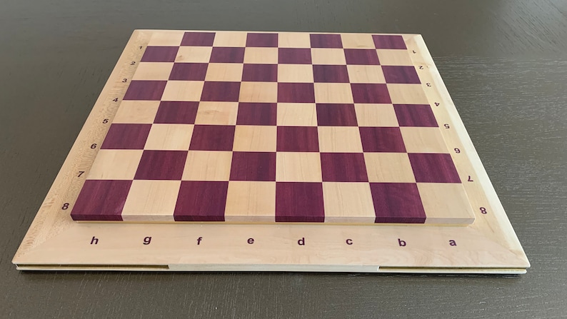 Puede incluir: Un tablero de ajedrez de madera con un patr&oacute;n de tablero de ajedrez de madera clara y madera p&uacute;rpura. El tablero tiene n&uacute;meros del 1 al 8 en el lateral y letras de la a a la h en la parte inferior.