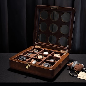 Puede incluir: Una caja de madera para relojes con tapa de cristal y 12 compartimentos para guardar relojes. La caja está abierta y hay relojes en algunos de los compartimentos. Hay un reloj y una pulsera en una pequeña almohada junto a la caja.