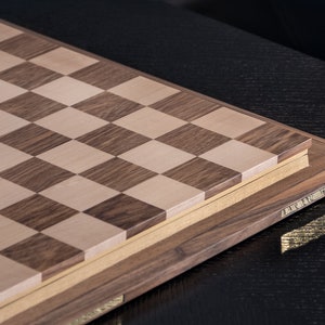 May include: A wooden chessboard with a light brown and dark brown chequered pattern. The board is closed and has a decorative border.