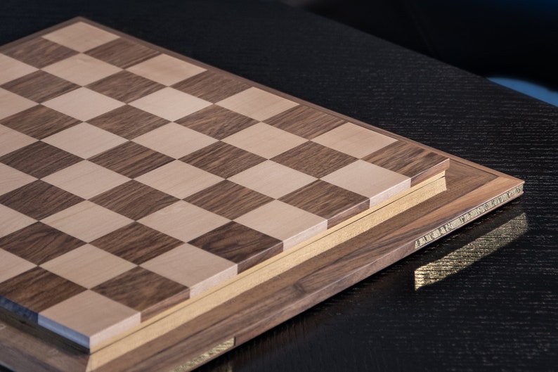 May include: A wooden chessboard with a light brown and dark brown chequered pattern. The board is closed and has a decorative border.