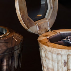 May include: Two wooden watch boxes, one open with a black watch inside and the other closed. The boxes are made of wood with a natural finish and have a unique design.