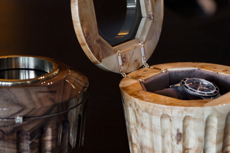 May include: Two wooden watch boxes, one open with a black watch inside and the other closed. The boxes are made of wood with a natural finish and have a unique design.