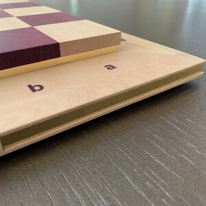 Puede incluir: Un tablero de ajedrez de madera con un patr&oacute;n de cuadros de madera p&uacute;rpura y claro. El tablero est&aacute; cerrado y tiene un borde de lat&oacute;n. El tablero est&aacute; etiquetado con las letras "a" y "b" en el lateral.