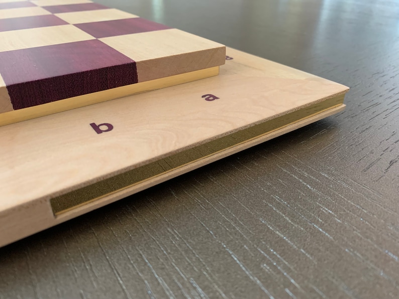 Puede incluir: Un tablero de ajedrez de madera con un patr&oacute;n de cuadros de madera p&uacute;rpura y claro. El tablero est&aacute; cerrado y tiene un borde de lat&oacute;n. El tablero est&aacute; etiquetado con las letras "a" y "b" en el lateral.