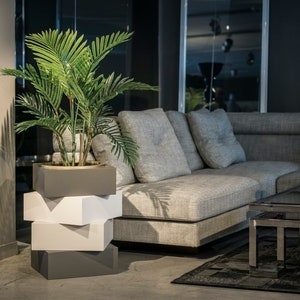 Puede incluir: Un sofá seccional gris con cojines grises y un soporte para plantas gris y blanco con una planta verde. El sofá está en una sala de estar moderna con una mesa de café de vidrio y una alfombra negra.