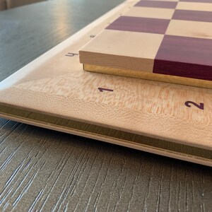 Puede incluir: Un tablero de ajedrez de madera con una base de madera clara y una superficie de juego de madera oscura. El tablero tiene un borde dorado y los n&uacute;meros 1 y 2 son visibles en la base.