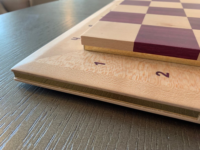 Puede incluir: Un tablero de ajedrez de madera con una base de madera clara y una superficie de juego de madera oscura. El tablero tiene un borde dorado y los n&uacute;meros 1 y 2 son visibles en la base.