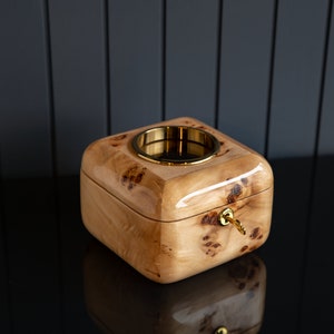 Puede incluir: Una caja de madera con una cerradura de latón y un anillo de latón en la parte superior. La caja está hecha de madera clara con un patrón marrón y blanco.