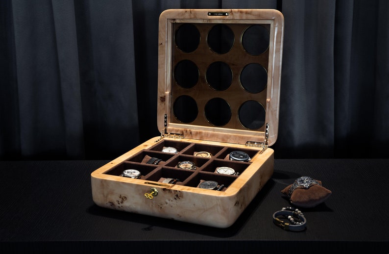 May include: A wooden watch box with a glass top and 12 compartments for storing watches. The box is made of light wood with a brown stain and has a gold-colored lock. The box is open and there are watches in some of the compartments.