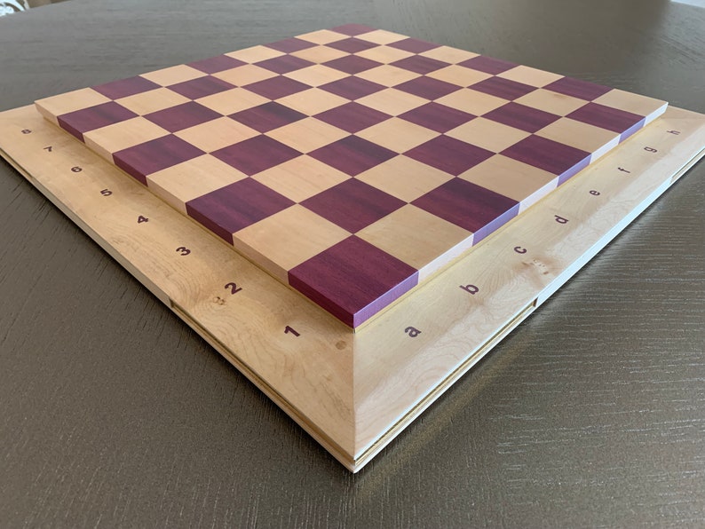 Puede incluir: Un tablero de ajedrez de madera con un patr&oacute;n de cuadros marr&oacute;n claro y morado. El tablero est&aacute; doblado por la mitad, mostrando los n&uacute;meros del 1 al 8 en un lado y las letras de la a a la h en el otro.