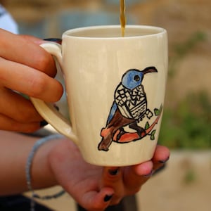 May include: Cream-colored ceramic mug with a hand-painted bird design. The bird features blue, brown, and black details, perched on a red branch with green leaves. The mug is being filled with a dark liquid.