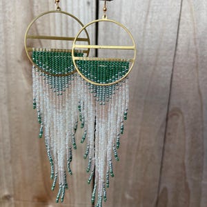May include: Gold-coloured hoop earrings with a half-circle of green beads and white beaded fringe. The earrings are suspended from a brown branch against a wooden backdrop. The earrings have a modern, geometric design.