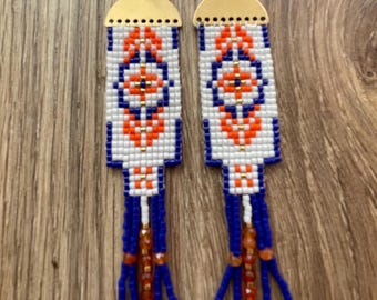Op de afbeelding: Een paar kralen oorbellen met een geometrisch patroon in oranje en blauw op een witte achtergrond. De oorbellen hebben goudkleurige metalen accenten en blauwe kralenfranjes. Gepresenteerd op een houten oppervlak.