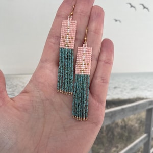 May include: Handmade beaded earrings with a rectangular design. The top section is pink with white and gold accents, transitioning to a turquoise fringe. Gold-colored ear wires complete the design. The earrings are held in a hand.