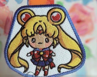 May include: A white embroidered patch featuring a cartoon Sailor Moon character with yellow hair, a red bow, and a blue sailor uniform. The patch has a blue border and is on a small wooden easel.