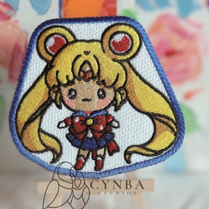 May include: A white embroidered patch featuring a cartoon Sailor Moon character with yellow hair, a red bow, and a blue sailor uniform. The patch has a blue border and is on a small wooden easel.