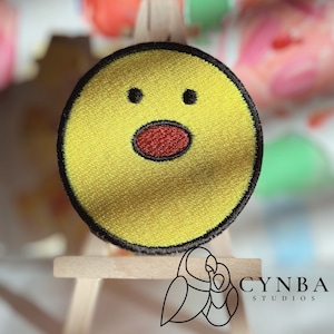 May include: Yellow felt circle with a black outline, depicting a surprised face with two black eyes and a red mouth. The circle is displayed on a small wooden easel.
