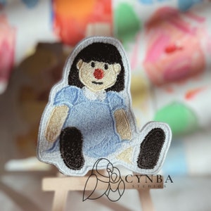 May include: A blue and white embroidered patch of a girl doll with black hair, a red nose, and a blue dress. The patch is sitting on a wooden easel.