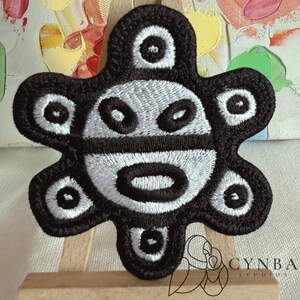 May include: Embroidered patch featuring a stylized black and white design. The patch has a central face-like motif with oval and circular shapes, surrounded by a petal-like border. The patch is likely for clothing or accessories.