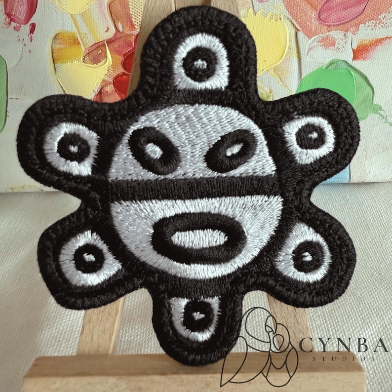 May include: Embroidered patch featuring a stylized black and white design. The patch has a central face-like motif with oval and circular shapes, surrounded by a petal-like border. The patch is likely for clothing or accessories.