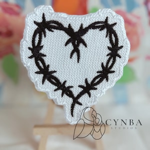 May include: White heart-shaped patch with a black barbed wire design. The patch is embroidered and has a slightly frayed edge.