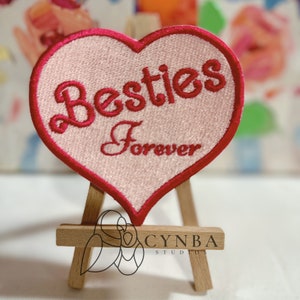 May include: Pink heart-shaped embroidered patch with red stitching. The patch says "Besties Forever" in a cursive font.