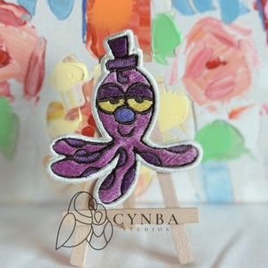 May include: Purple octopus with a top hat, embroidered patch. The octopus has a blue nose and yellow eyes. The patch is on a small wooden easel.