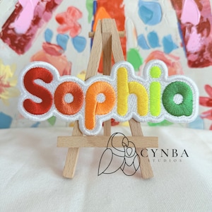 May include: A colorful embroidered patch with the name "Sophia" in rainbow colors. The patch is on a small wooden easel.
