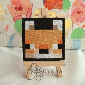 May include: A square embroidered patch featuring a pixelated fox face design in shades of orange, white, black, and tan, framed by a thick black border. The patch is displayed on a small wooden easel, with the text CYNBA STUDIOS visible below.