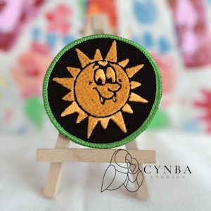 May include: A round embroidered patch with a green border. The patch features a cartoon sun with a toothy grin and large eyes. The sun is orange and the background is black.