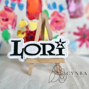 May include: White embroidered patch with the name "Lori" in black lettering and a black star design. The patch is displayed on a small wooden easel.